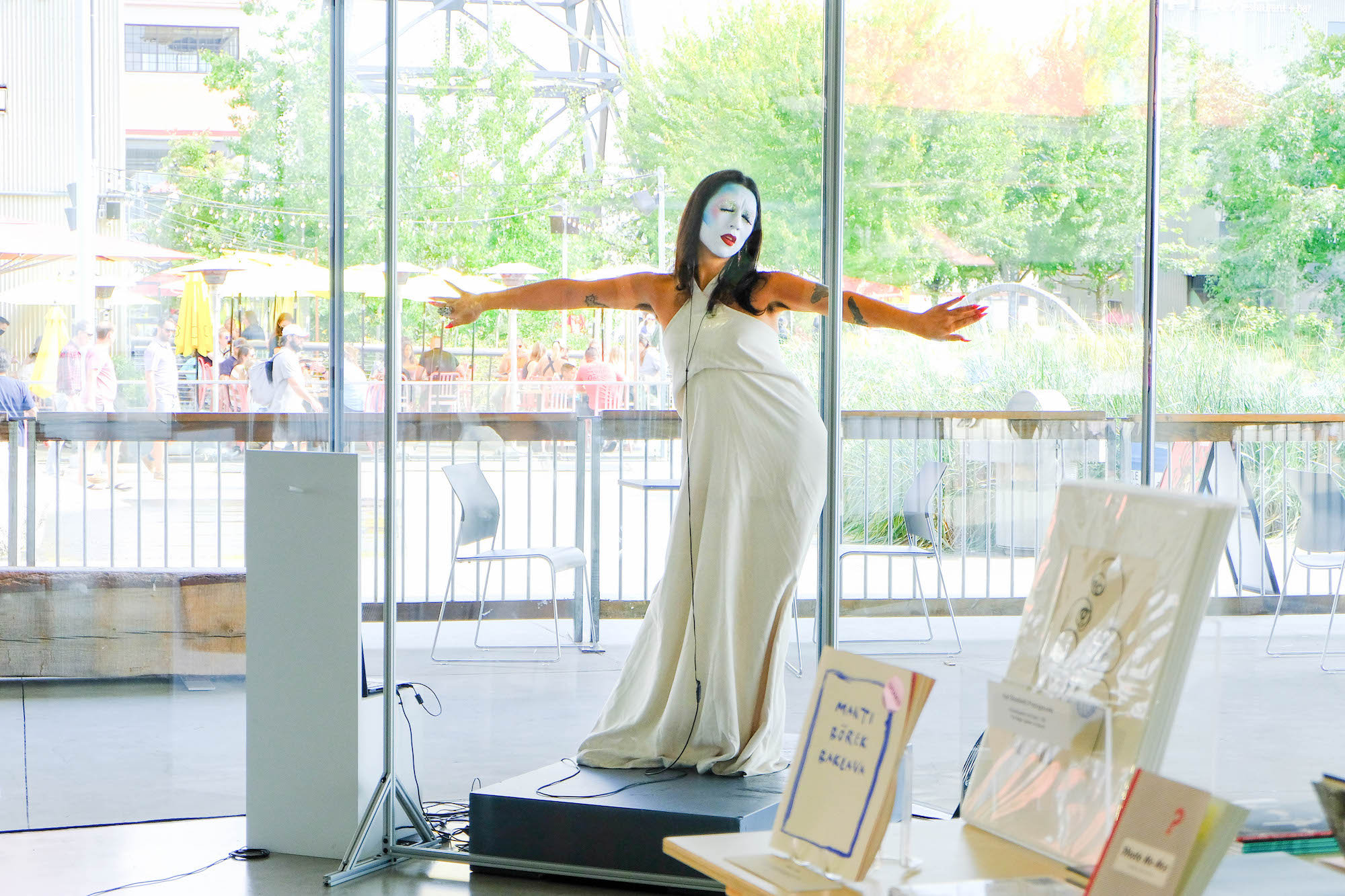 Inside a gallery space, with big glass windows behind them, House of Rice member Maiden China, with full white and blue face make-up, performs with eyes closed and arms spread out.