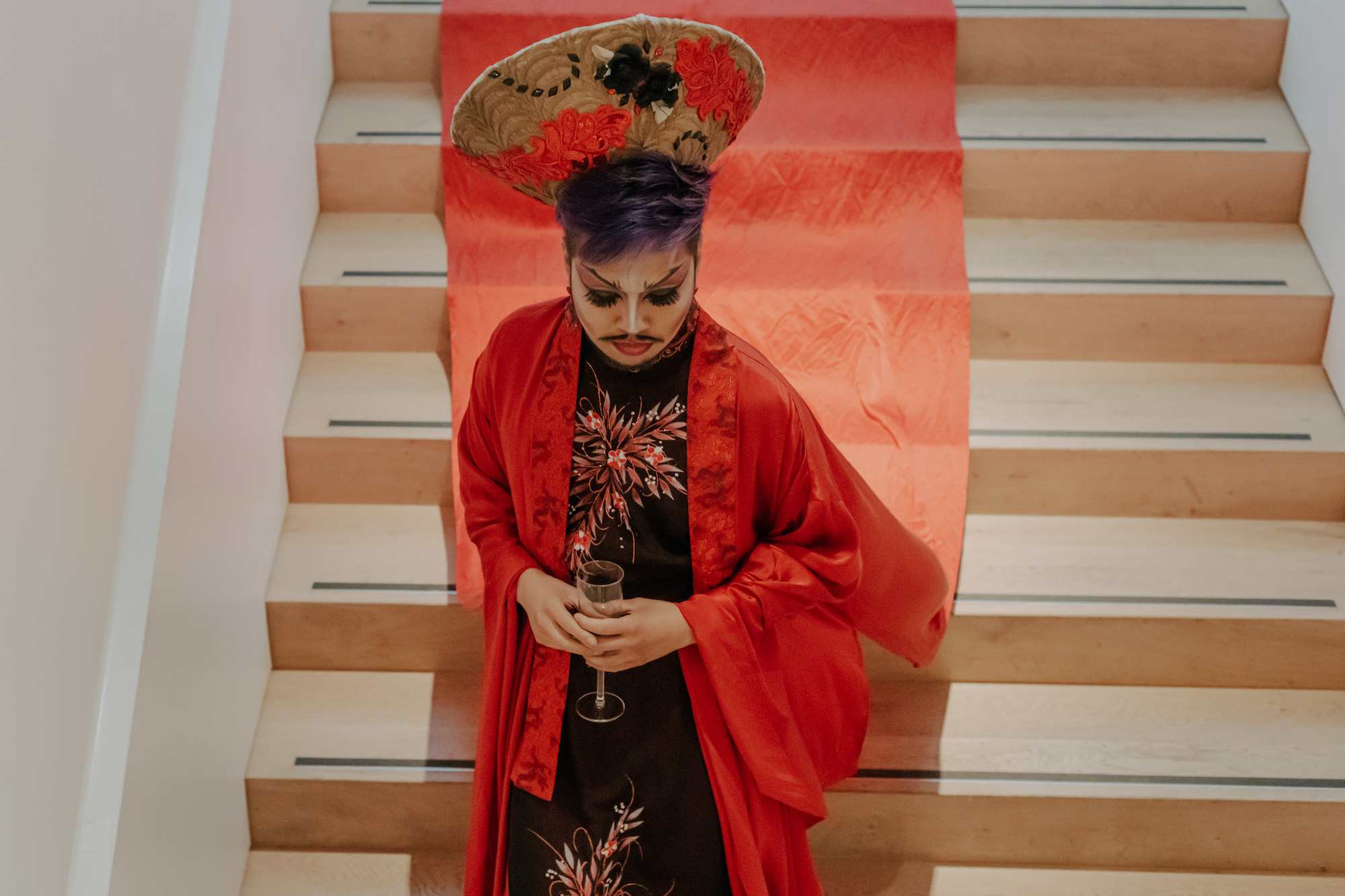 Seen from above, House of Rice member Shay Dior, wearing an embroidered hat, a black cheongsam, and a red robe, descends a staircase, with the red robe flowing behind them.