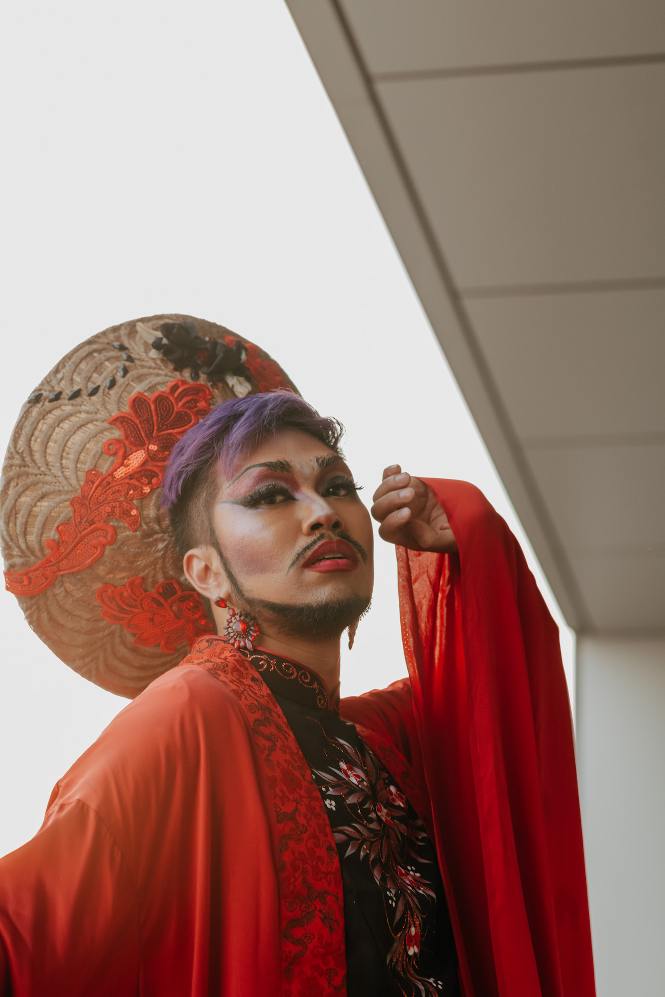 House of Rice member Shay Dior, wearing a an embroidered hat, a black choengsam, and a red robe, poses with their hand held near their face.