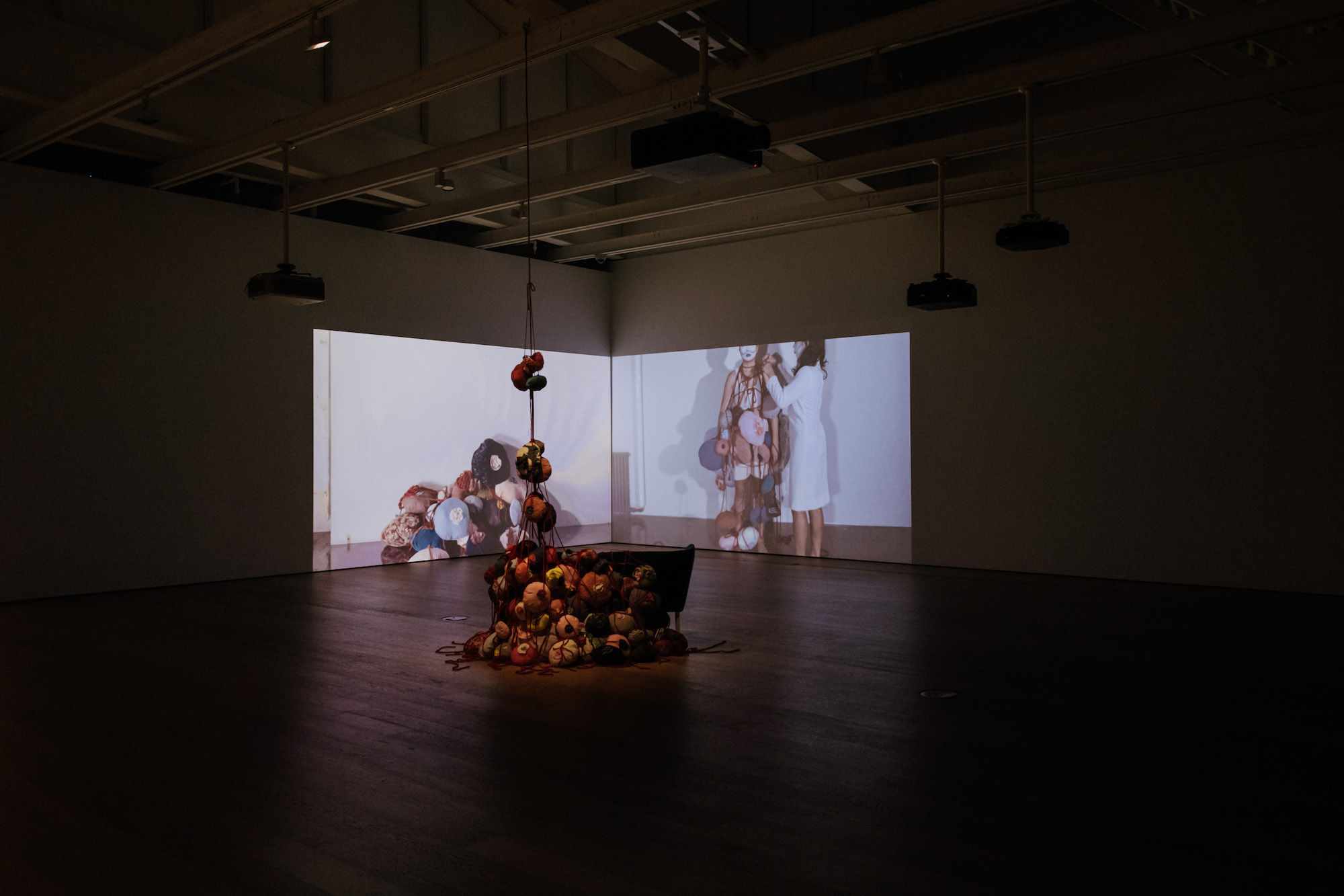 An installation image of a two-channel video, on the left is a pile of bulbous shapes made of fabric. On the right, a House of Rice member has these objects draped on them by a second person. In front of the screen, in the middle of the room, we see the same bulbous objects in a pile, suspended from the ceiling.