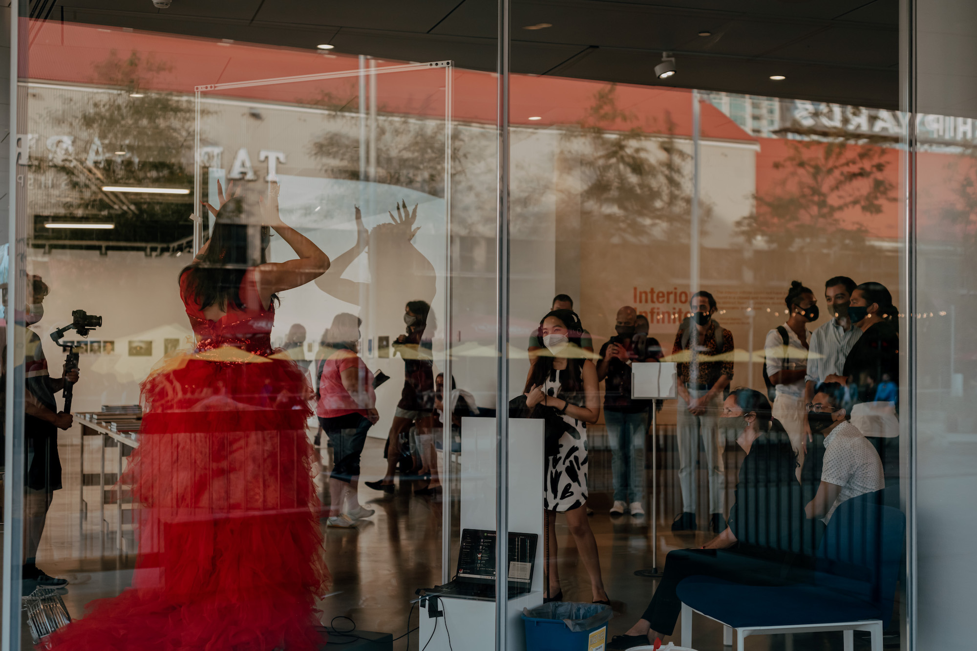 Seen through a window, House of Rice member Maiden China, dressed in a red dress, performs for an audience with her open hands raised to her temples.