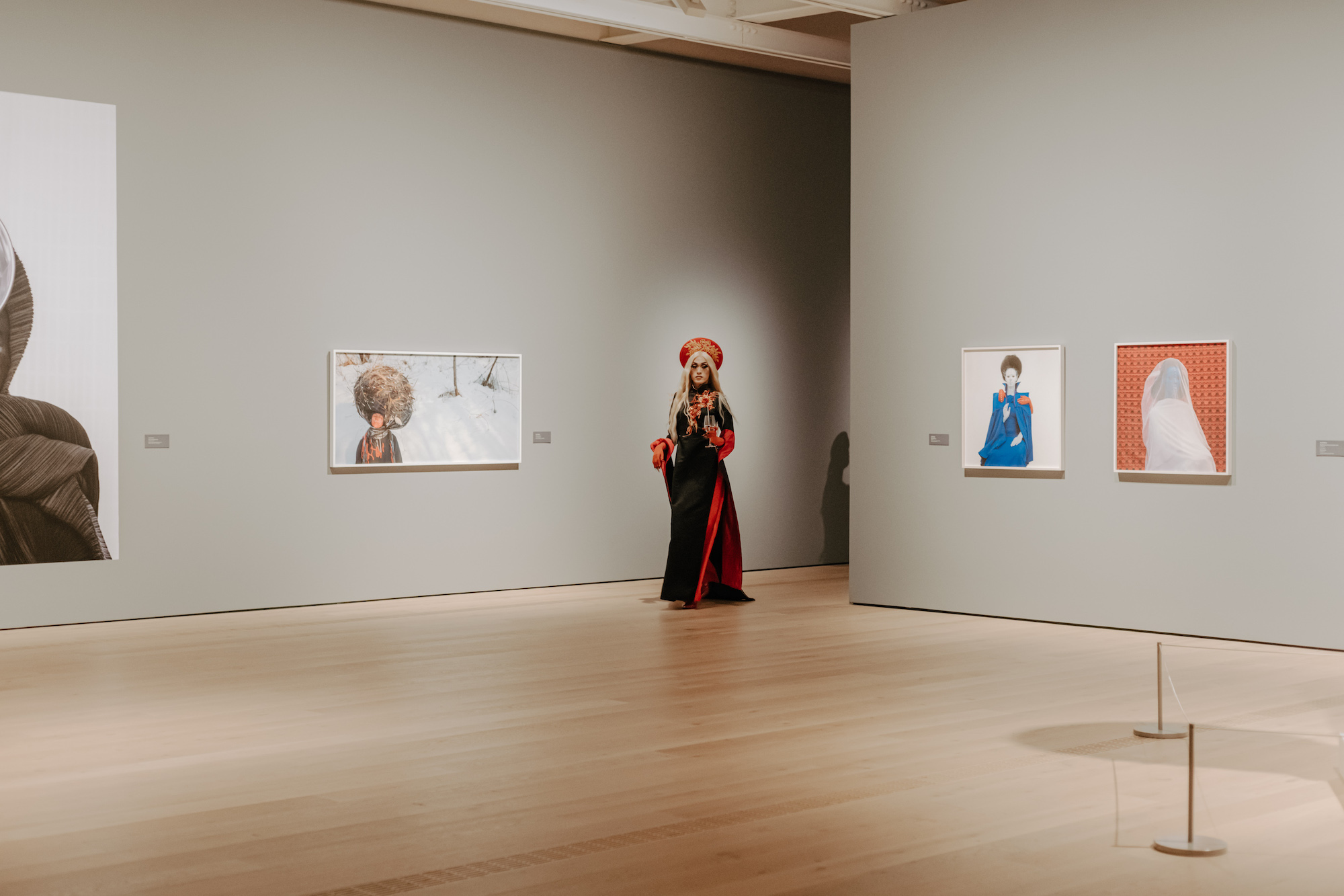 A wide composition in the gallery, with House of Rice member Kara Juku dressed in a black and red cheongsam. To their left is a photo by Meryl McMaster.