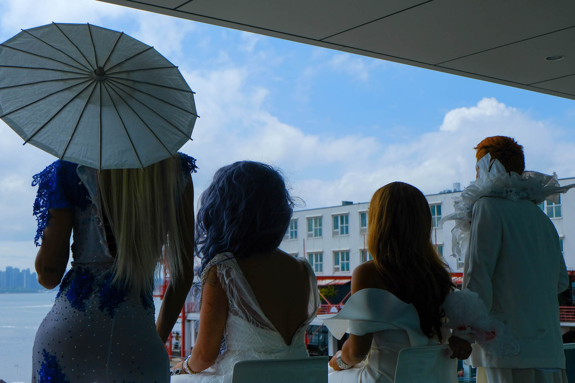 Both seated and standing on the Polygon Gallery balcony, four members of House of Rice dressed in white are seen from behind as they overlook the water and a distant Vancouver skyline. One holds a parasol.