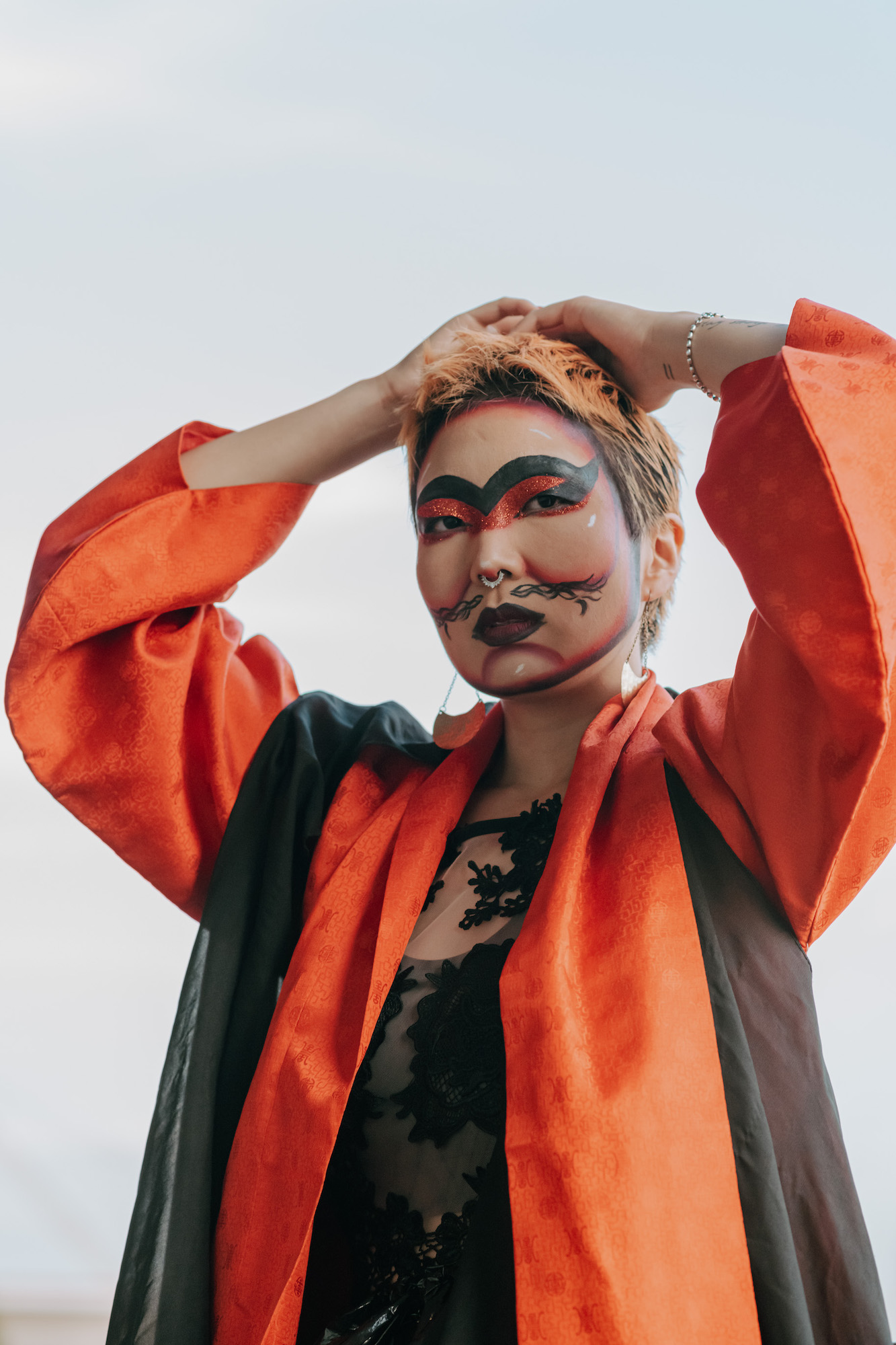 House of Rice member Skim, with orange hair and bold make-up and wearing an orange and black robe, poses with his hands above his head.