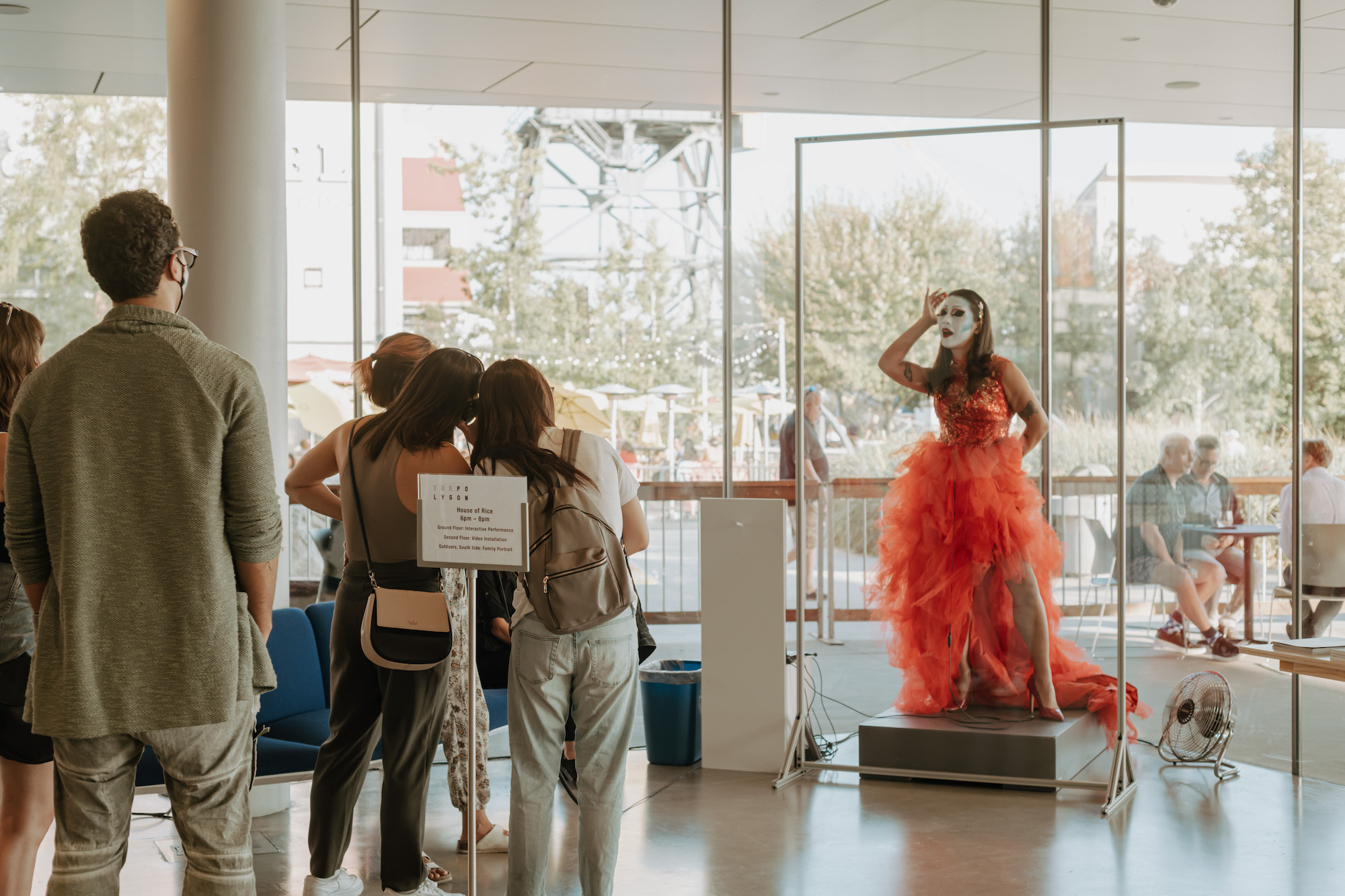 Inside a gallery space, with big glass windows behind them, House of Rice member Maiden China, with full white make-up and a red dress, performs with her eyes closed and her arm raised in front of an audience.