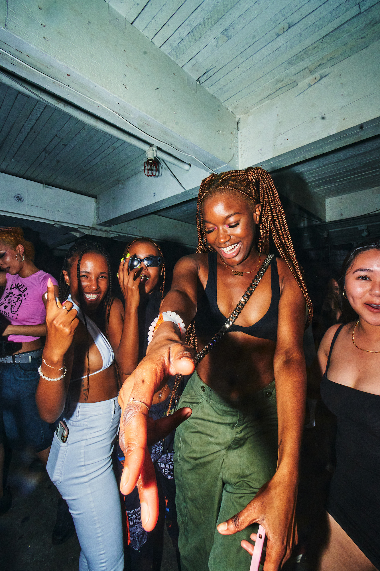 A wide-angle vertical photo of a Black woman dancing and smiling with her hand stretched towards the camera. She is surrounded by people on her left and right who are also smiling and looking at the camera.