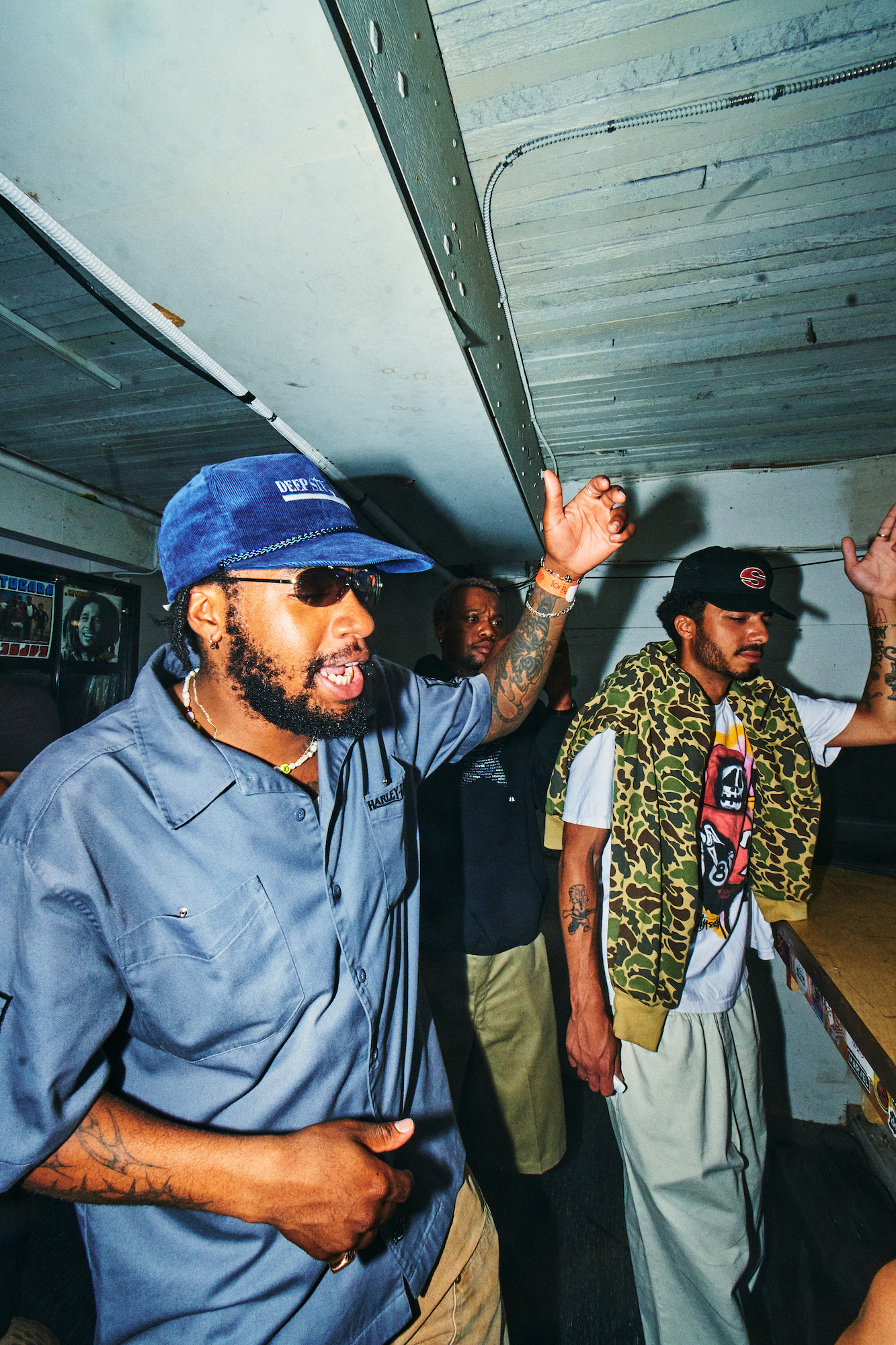 A Black man in a blue shirt, a blue baseball cap, and sunglasses sings along to a song with his arm raised, beside two other men.