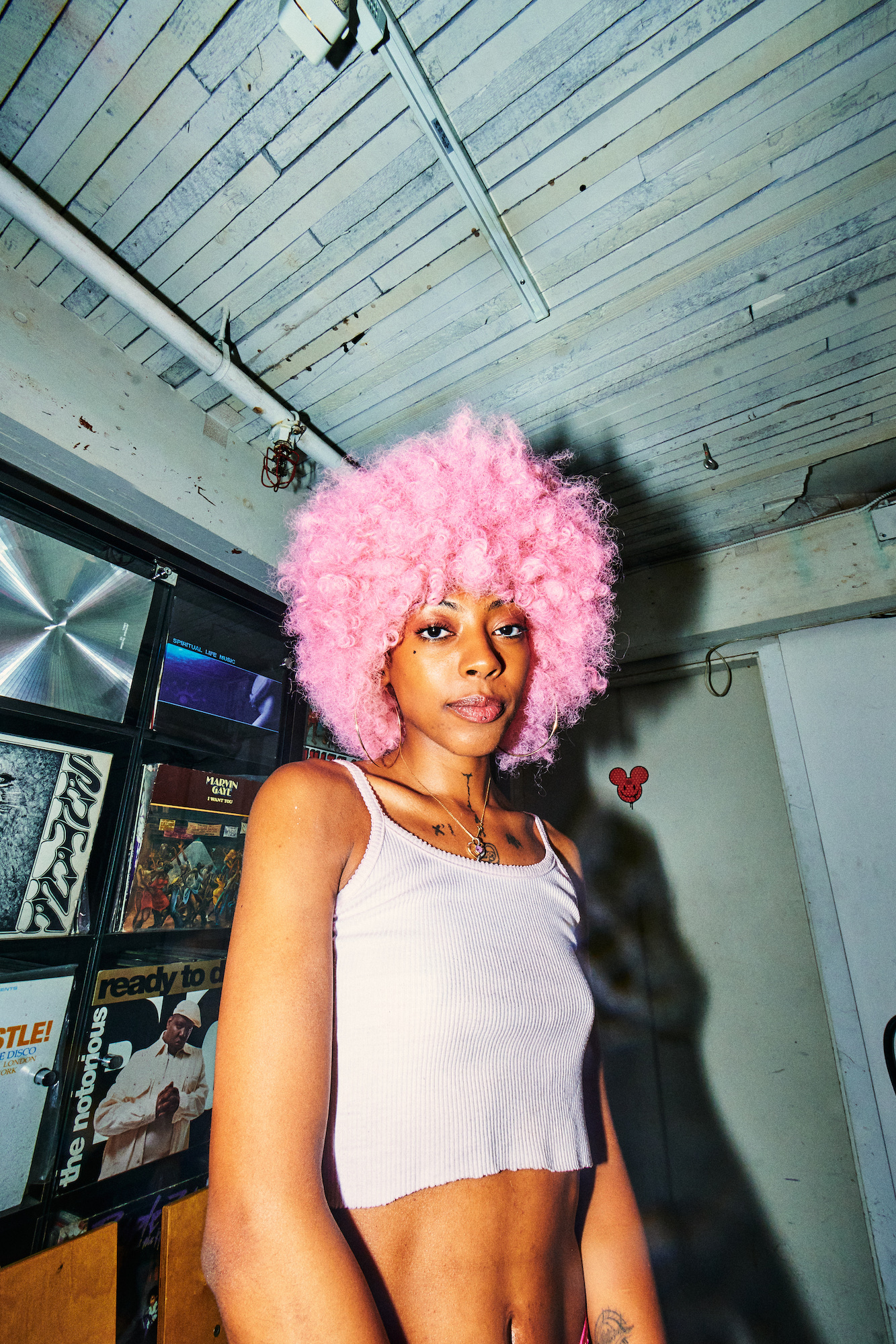 A low wide-angle shot of a Black woman in a pink afro wig and a cropped white ribbed tank top looking right at the camera. The back wall displays vintage vinyls.