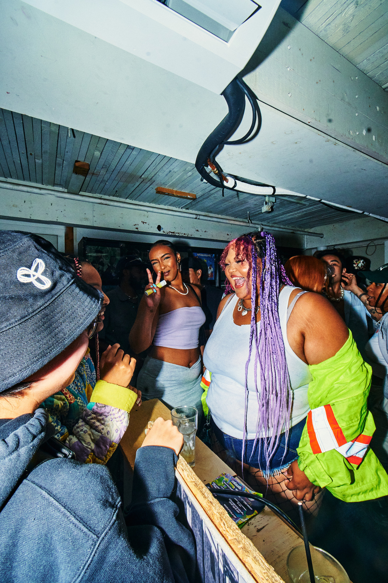 A party scene. In the centre a Black woman with purple hair smiles wide, beside another Black woman who flashes a peace sign to the camera.