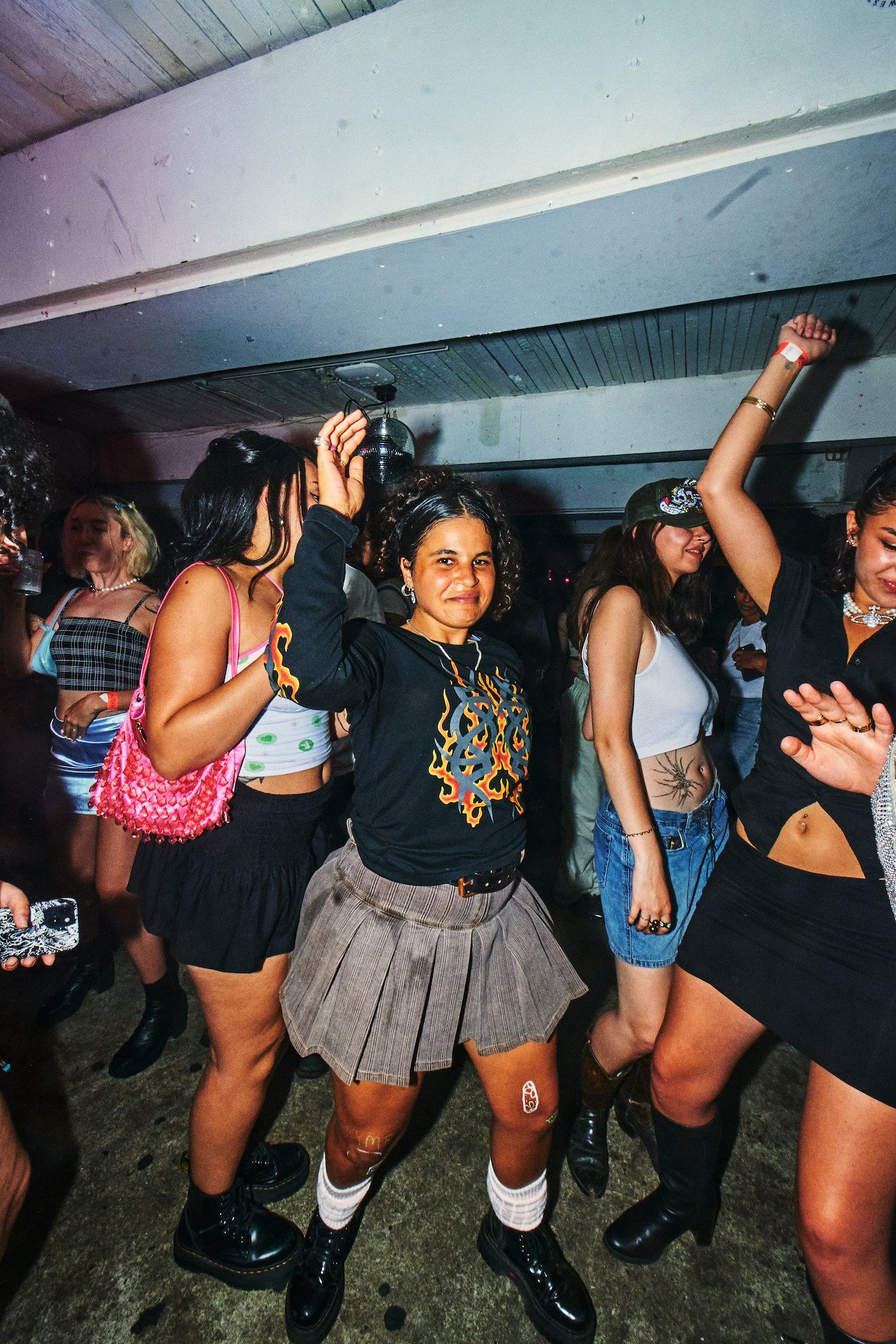 A crowd of people dance with their hands in the air. One woman in the middle, wearing a graphic t-shirt and a skirt, smiles to the camera.